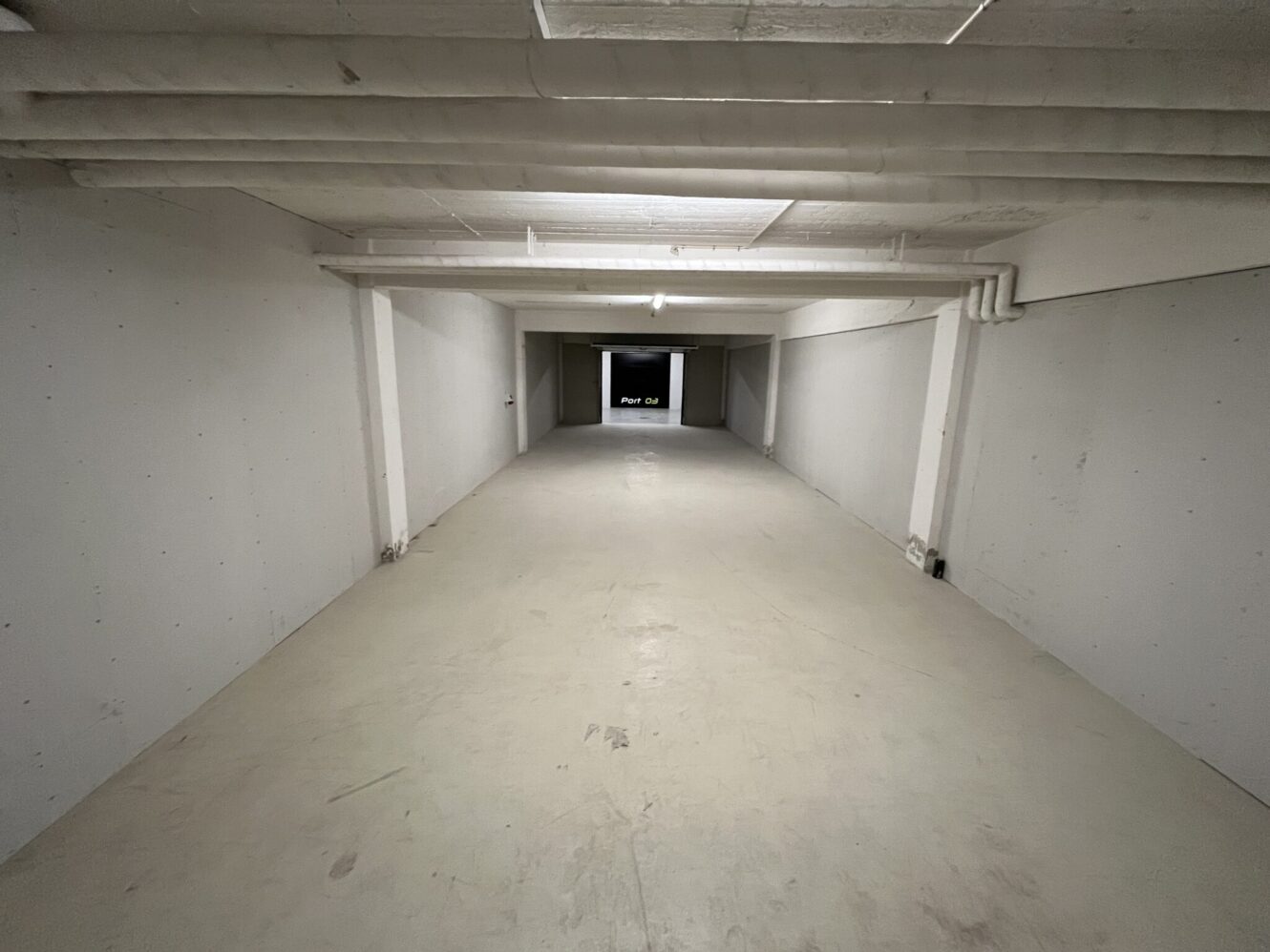 En lang, tom kældergang med hvide vægge, synlige rør i loftet og en dør i den anden ende.