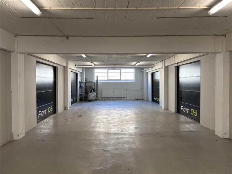 En ren, tom garage med fire individuelle sorte døre mærket Port 06 og Port 03 på hver side, betongulv og et vindue på bagsiden, der lukker naturligt lys ind.