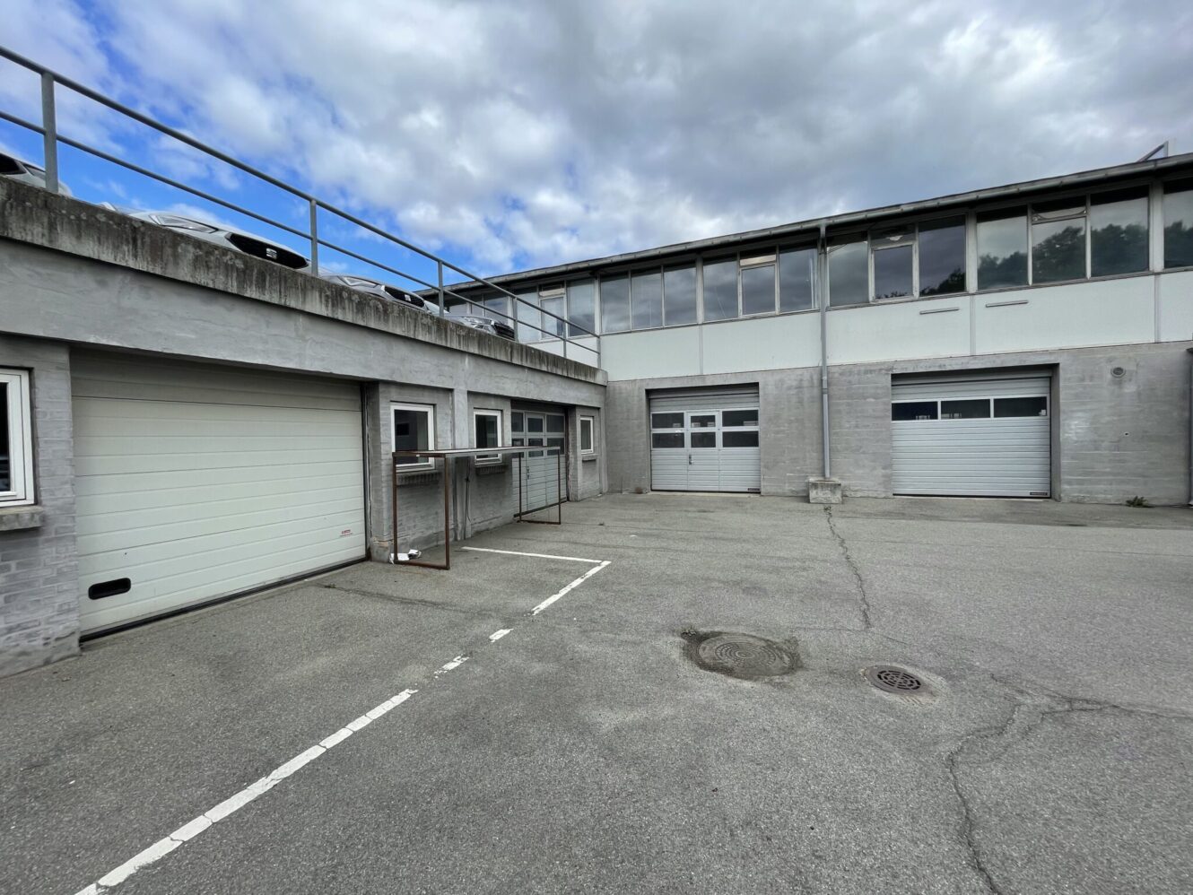 En industribygning i beton med tre store garageporte, små vinduer og et indhegnet område på en tom asfalteret grund under en overskyet himmel.