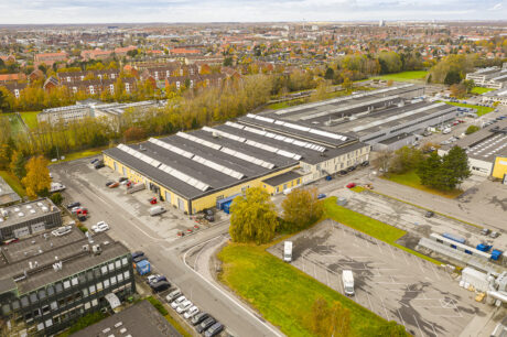 Luftfoto af et stort industrielt lagerkompleks med parkerede køretøjer, omgivet af træer og beboelsesbygninger i baggrunden.