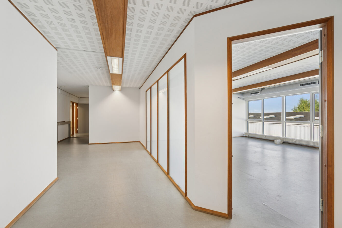 En ren, tom gang med hvide vægge, træbeklædning, matterede glaspaneler og en åben dør, der fører ind til et lyst rum med store vinduer.