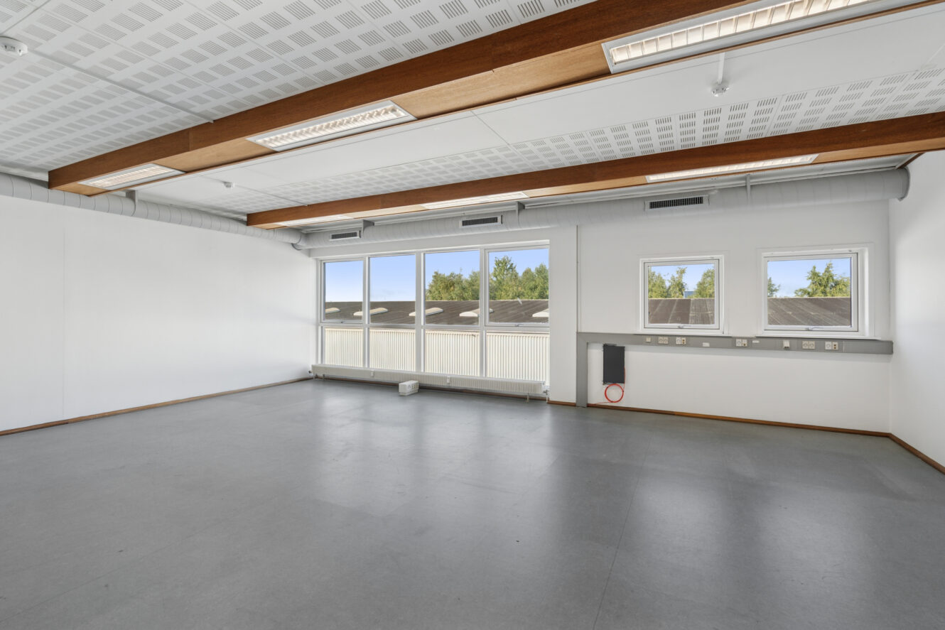 Tomt kontorlokale med hvide vægge, store vinduer, gråt gulv og synlige loftsbjælker. Naturligt lys kommer ind gennem vinduerne med udsigt til træer og hustage.
