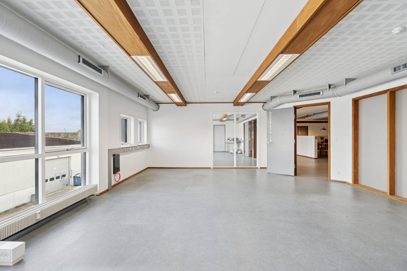 Et lyst, tomt kontorlokale med store vinduer, synlige kanaler, træbjælker i loftet og lysegråt gulv. Et tilstødende rum er synligt gennem en åben døråbning.