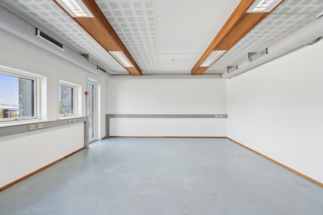 Tomt, moderne rum med hvide vægge, blågråt gulv, store vinduer, loftsventiler, synlige bjælker og fluorescerende belysning.