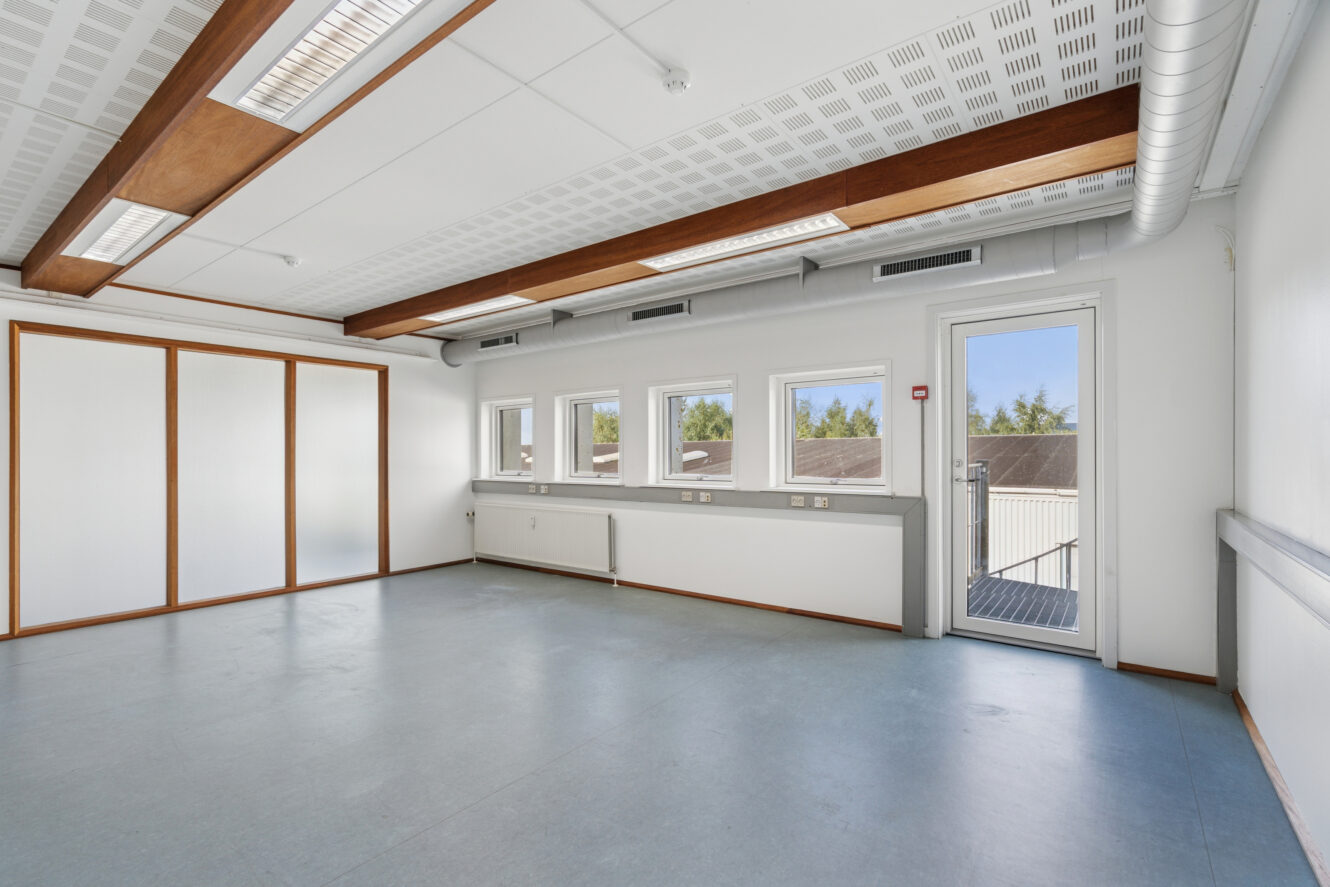 Et lyst, tomt rum med blåt gulv, hvide vægge, flere vinduer, en glasdør og træbjælker i loftet.