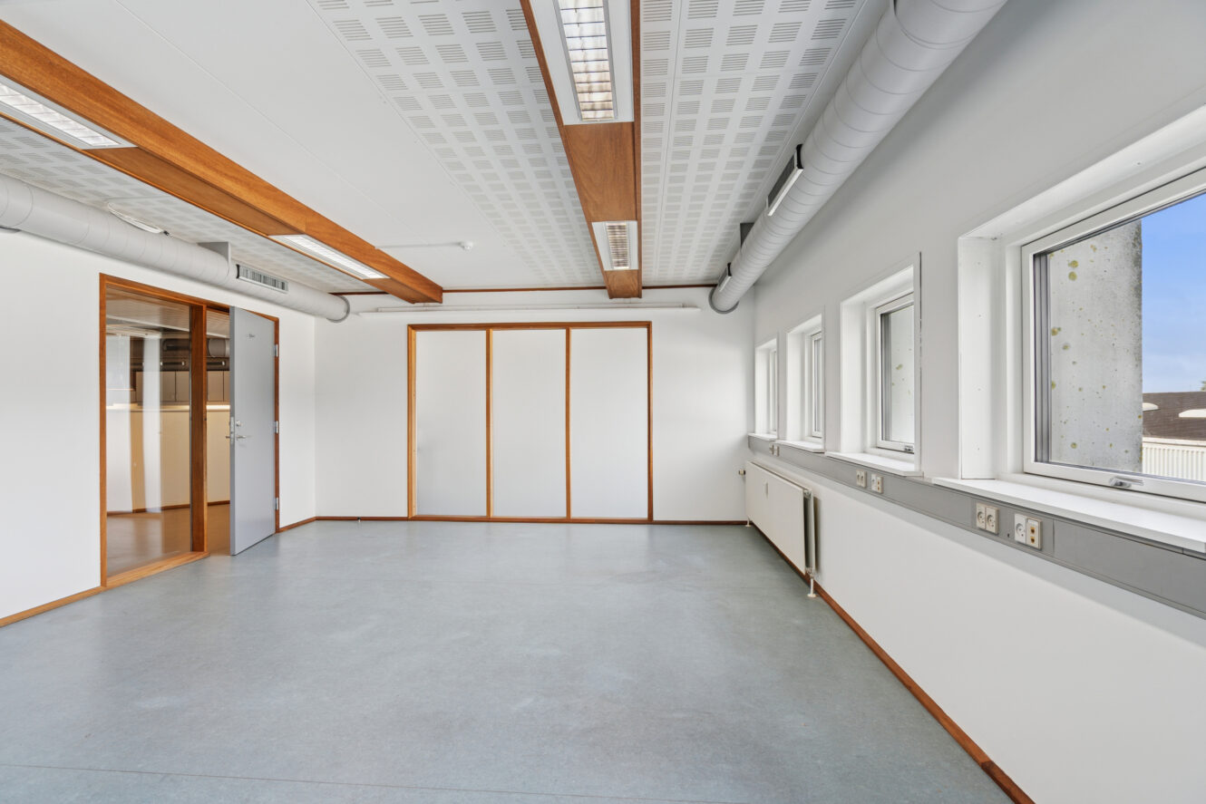 Tomt kontorlokale med hvide vægge, store vinduer, gråt gulv, træbjælker i loftet og lysstofrør.