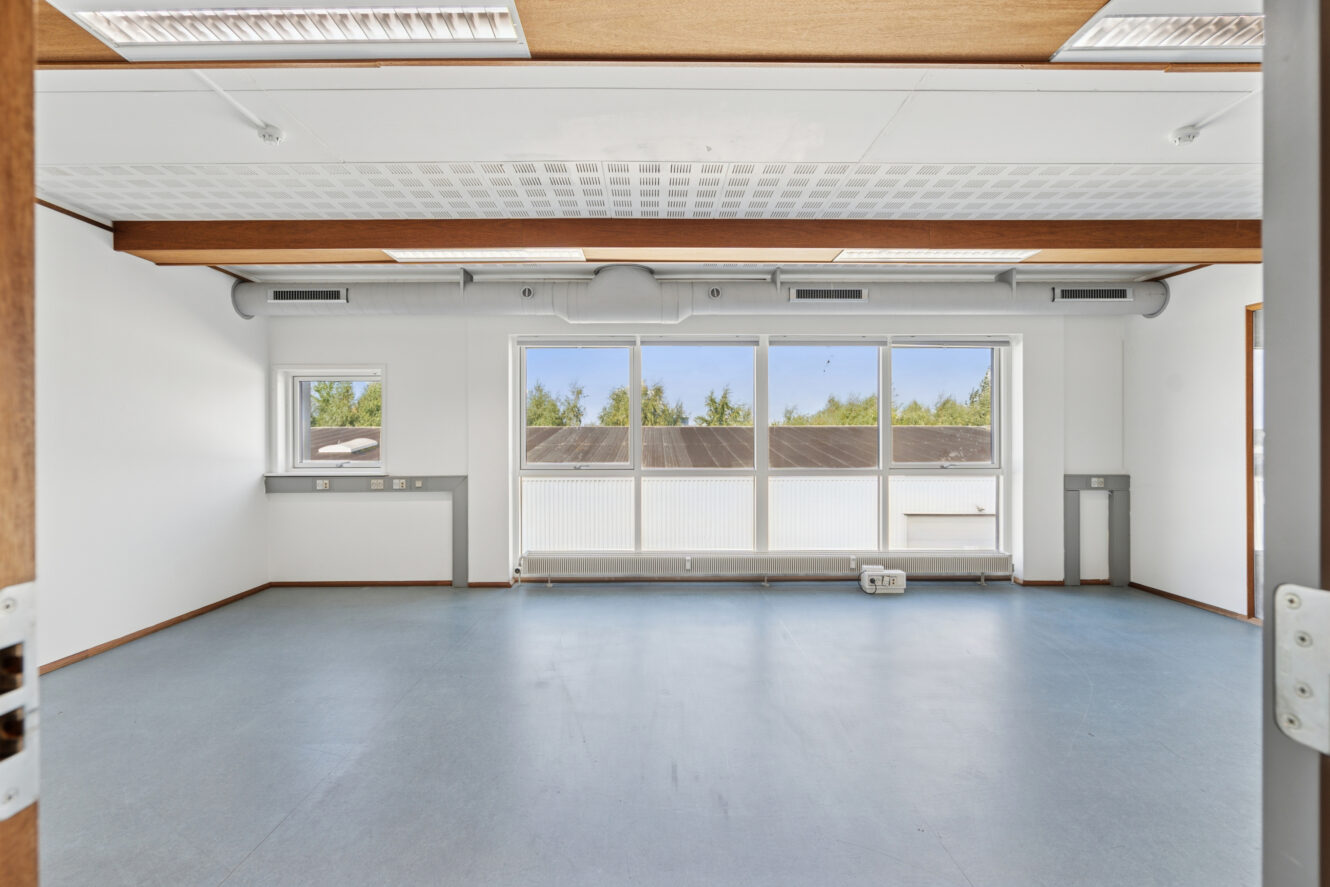 Tomt, umøbleret rum med blåt gulv, hvide vægge, store vinduer og synlige loftsbjælker; naturligt lys kommer ind gennem vinduerne.