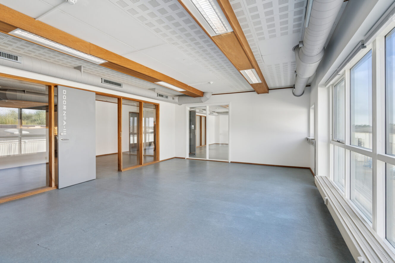 Tomme, moderne kontorlokaler med store vinduer, glasdøre, hvide vægge, blågråt gulv og synlige loftsdetaljer.