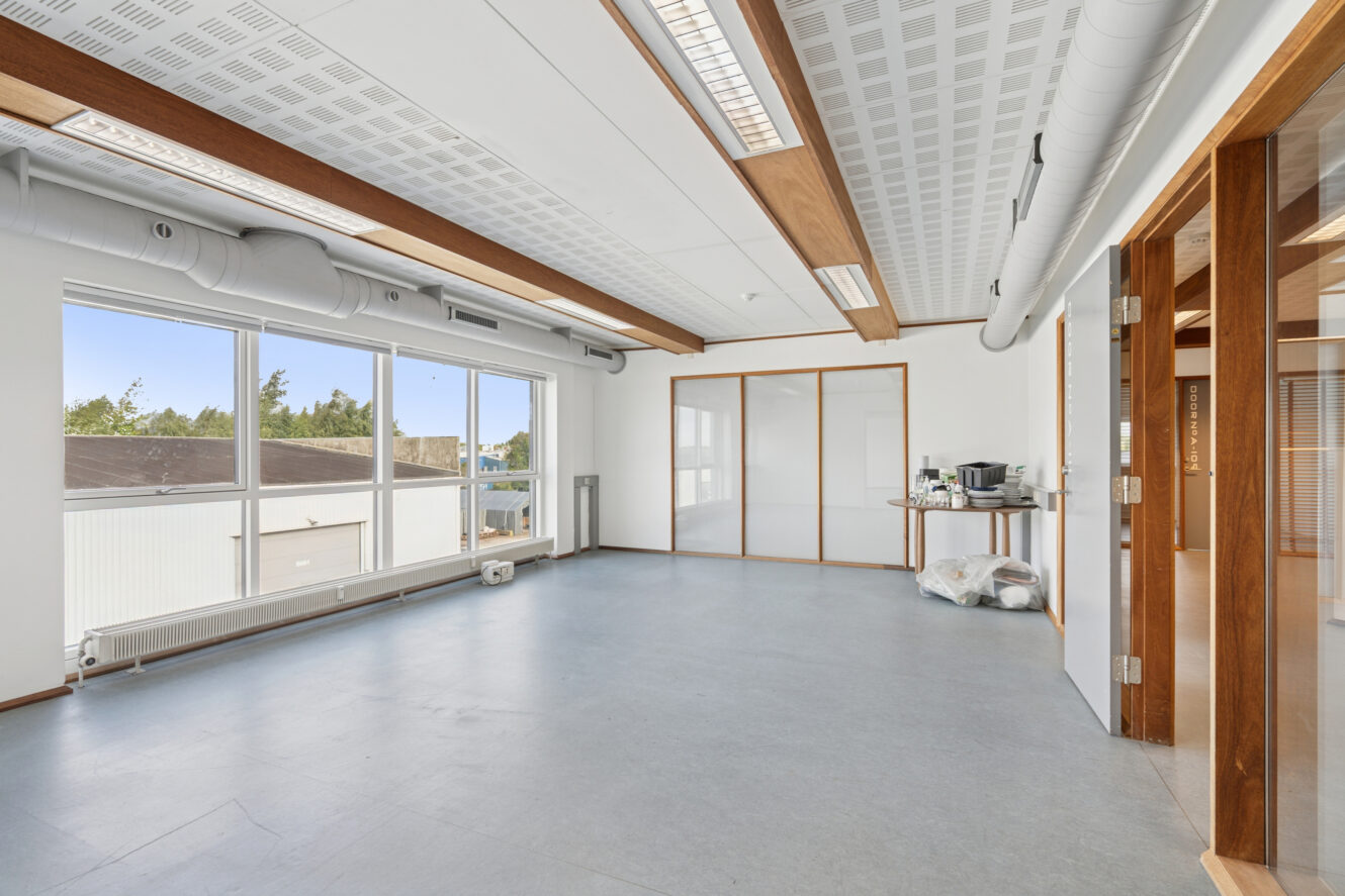 Tomt kontorlokale med store vinduer, synlige loftskanaler, trædetaljer og en kaffemaskine på et bord i hjørnet. Naturligt lys fylder rummet.