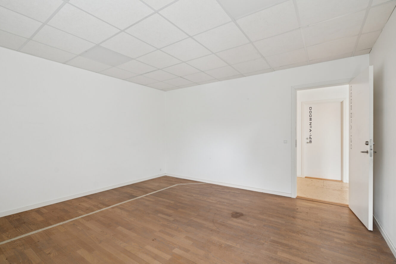 Tomt rum med hvide vægge, trægulv, hvidt loft med forsænkede paneler og en åben dør, der fører ud til en gang med lignende gulvbelægning.