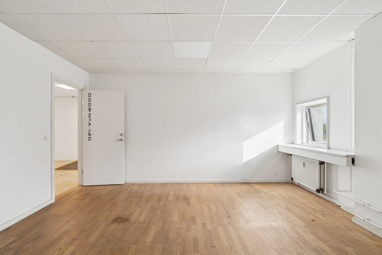 Et tomt, hvidmuret rum med trægulv, en lukket hvid dør til venstre og et vindue med radiator og rør på højre væg.