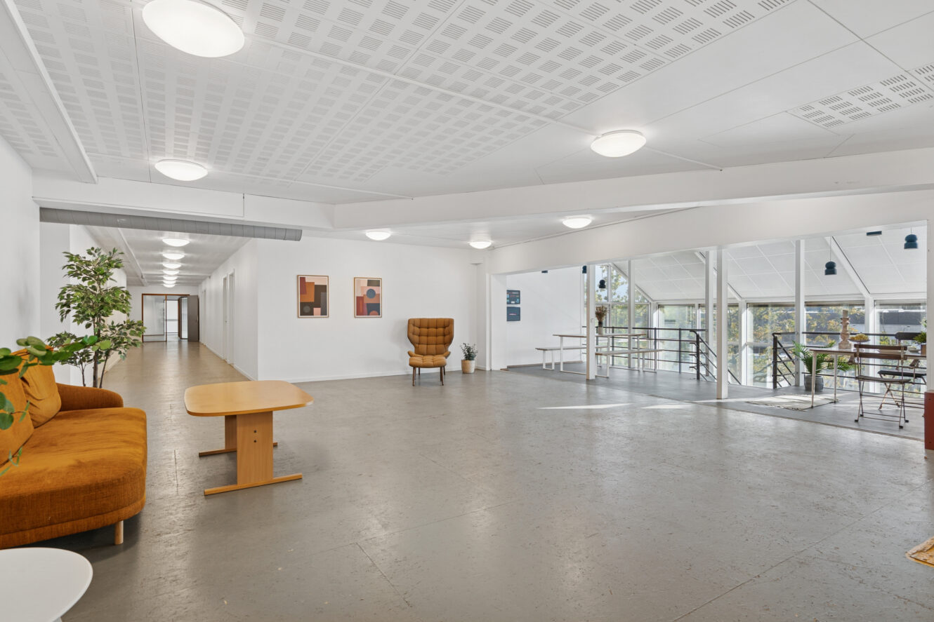 Moderne, rummeligt loungeområde med brune sofaer, et træbord, potteplanter, vægkunst og store vinduer med udsigt til et udendørs område; gangen ses til venstre.