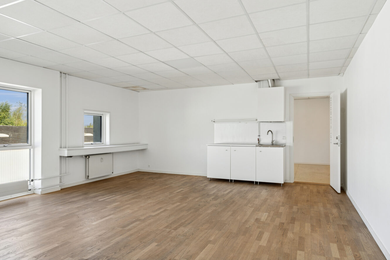 Et tomt rum med hvide vægge, trægulv, et lille tekøkken, to vinduer og en dør, der fører til et andet område.