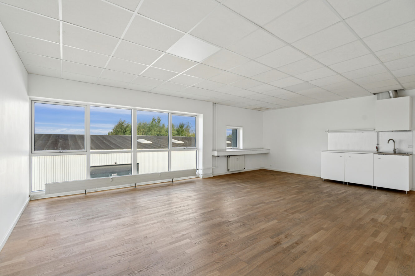 Tomt rum med trægulv, hvide vægge, store vinduer og et lille tekøkken med vask og skabe.