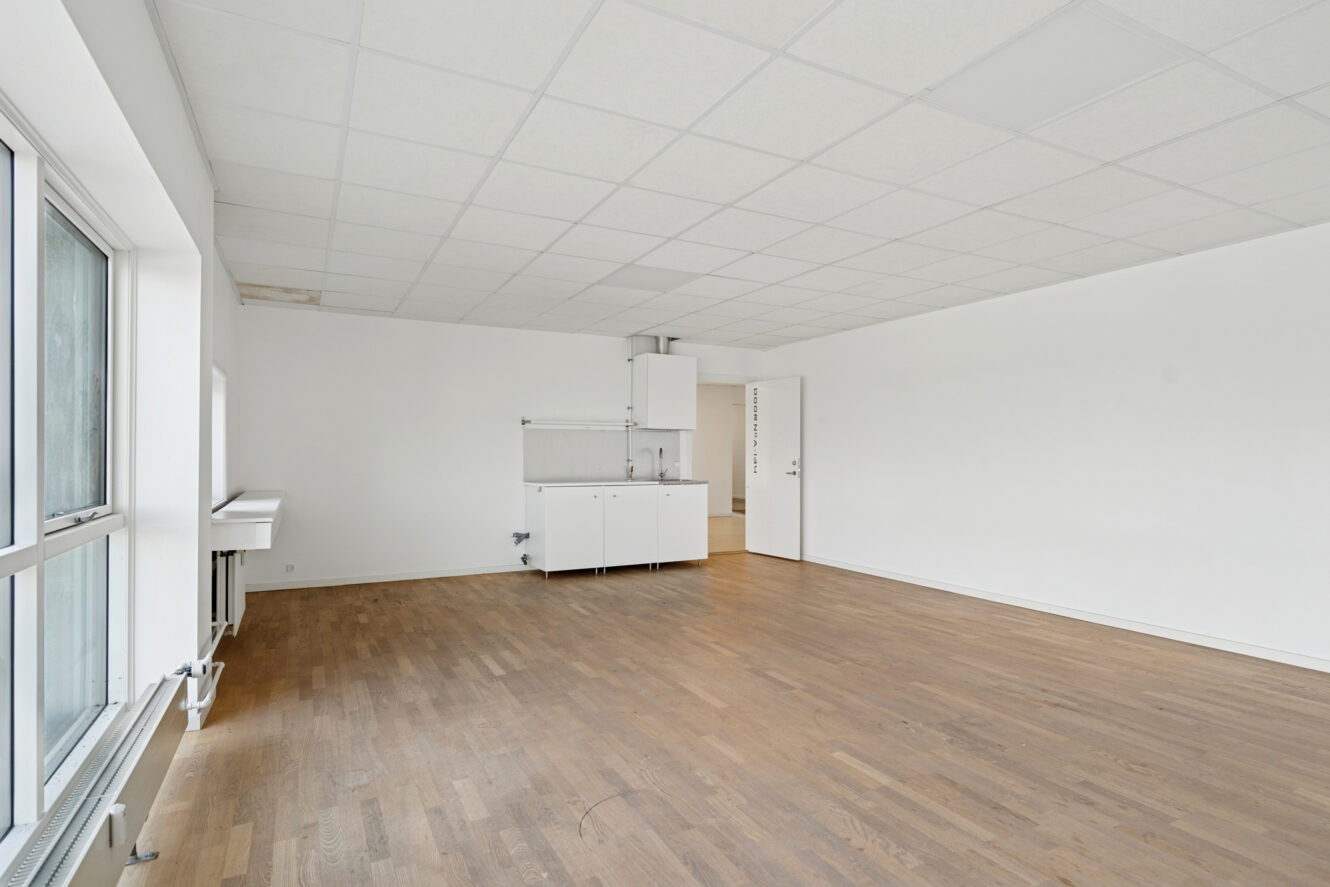 Umøbleret værelse med hvide vægge, store vinduer, trægulv og et lille tekøkken i det ene hjørne.