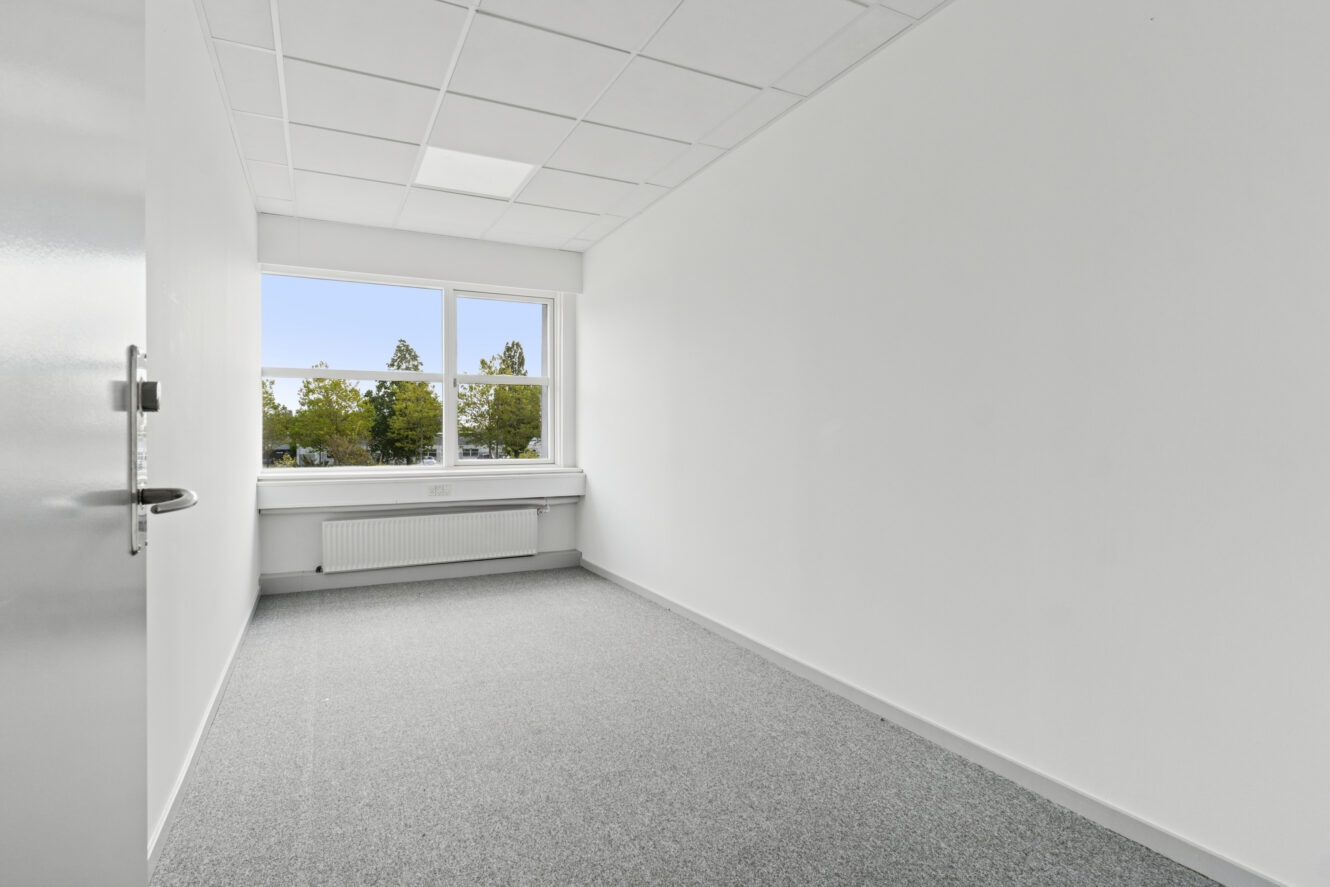 Lille tomt kontorlokale med hvide vægge, et vindue med udsigt til træer, gråt gulvtæppe og en dør, der står lidt åben.