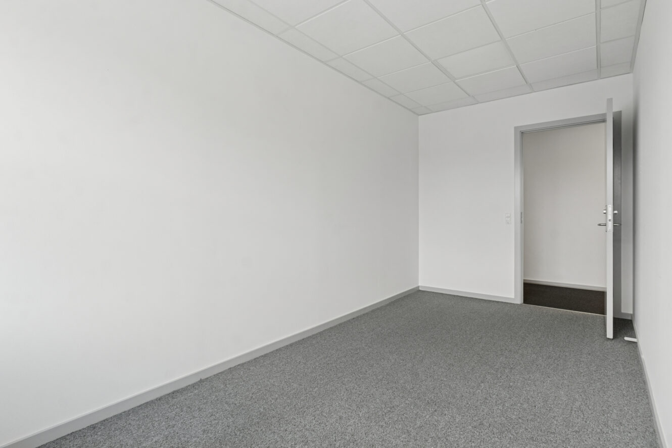 Tomt kontorlokale med hvide vægge, gråt gulvtæppe og en delvist åben dør, der fører til et andet rum. Loftet har hvide gitterpaneler.