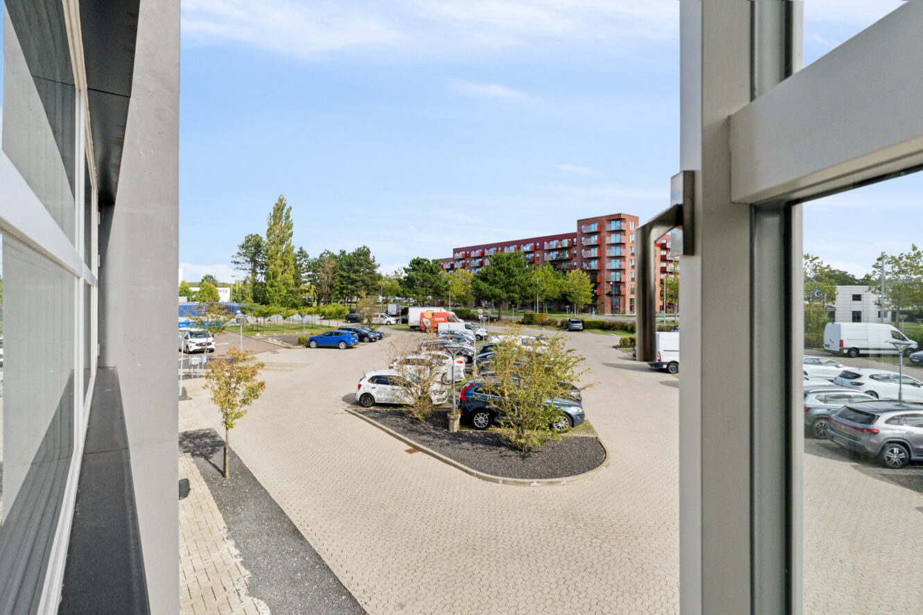 Udsigt fra et vindue over en parkeringsplads med flere biler, træer og en rød lejlighedsbygning i baggrunden under en blå himmel.