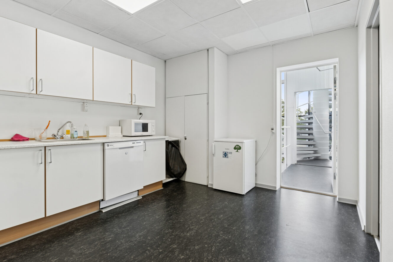 Lille kontorkøkken med hvide skabe, opvaskemaskine, mikroovn og minikøleskab; rengøringsartikler på køkkenbordet. Døråbningen fører til en trappeopgang med naturligt lys.