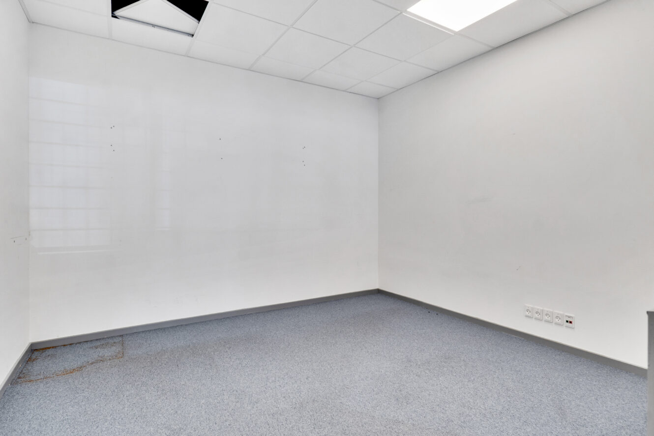Et tomt, umøbleret kontorlokale med hvide vægge, et blåt gulvtæppe, loftplader, nogle synlige vægpletter og et beskadiget loftpanel.