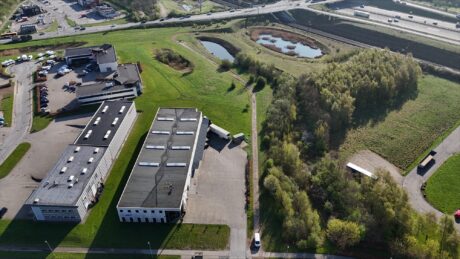 Luftfoto af industribygninger, parkeringsplads, veje og grønne områder med to damme og træer ved siden af en motorvej.