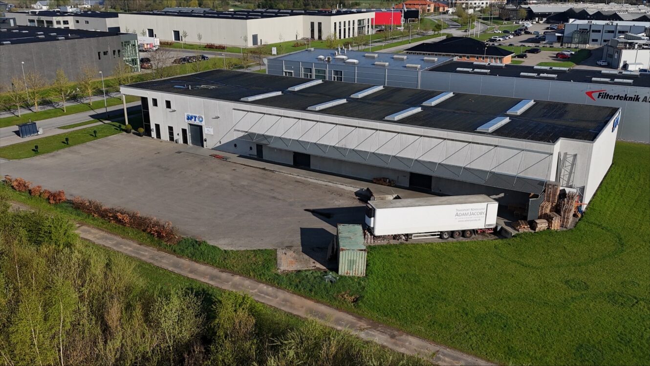 Luftfoto af en lagerbygning med en parkeret lastbil ved læsserampen, omgivet af græs og tilstødende industrianlæg.