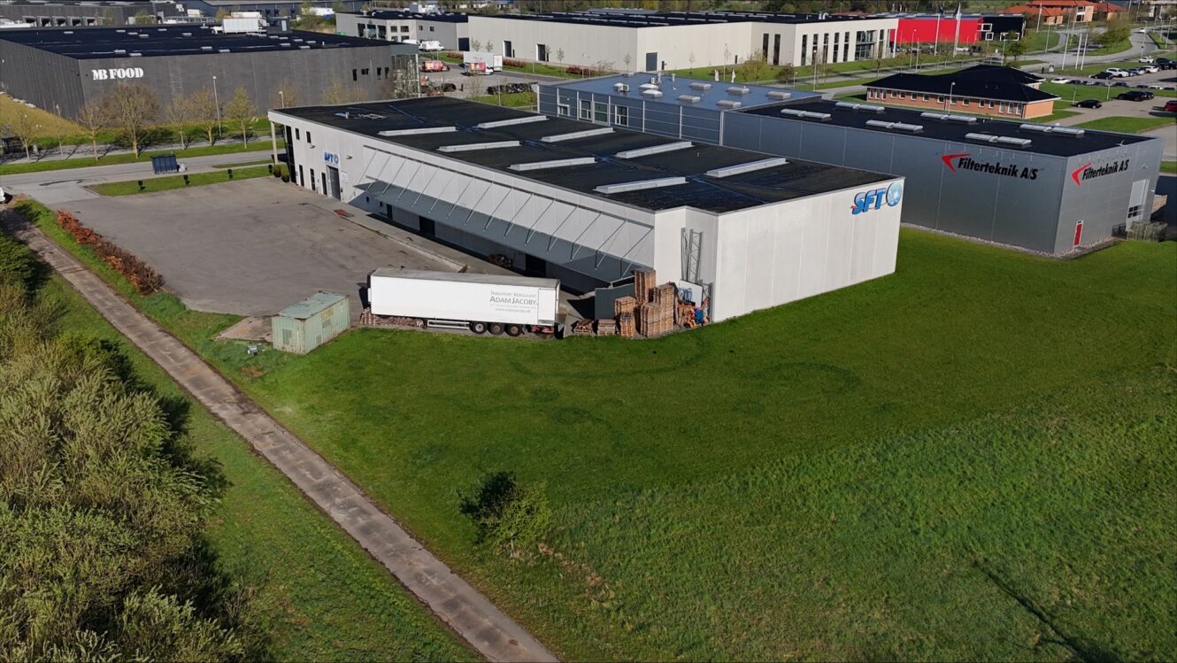 Luftfoto af en industribygning med en parkeret hvid sættevogn og stablede paller ved siden af, omgivet af græs og nærliggende lagerbygninger.