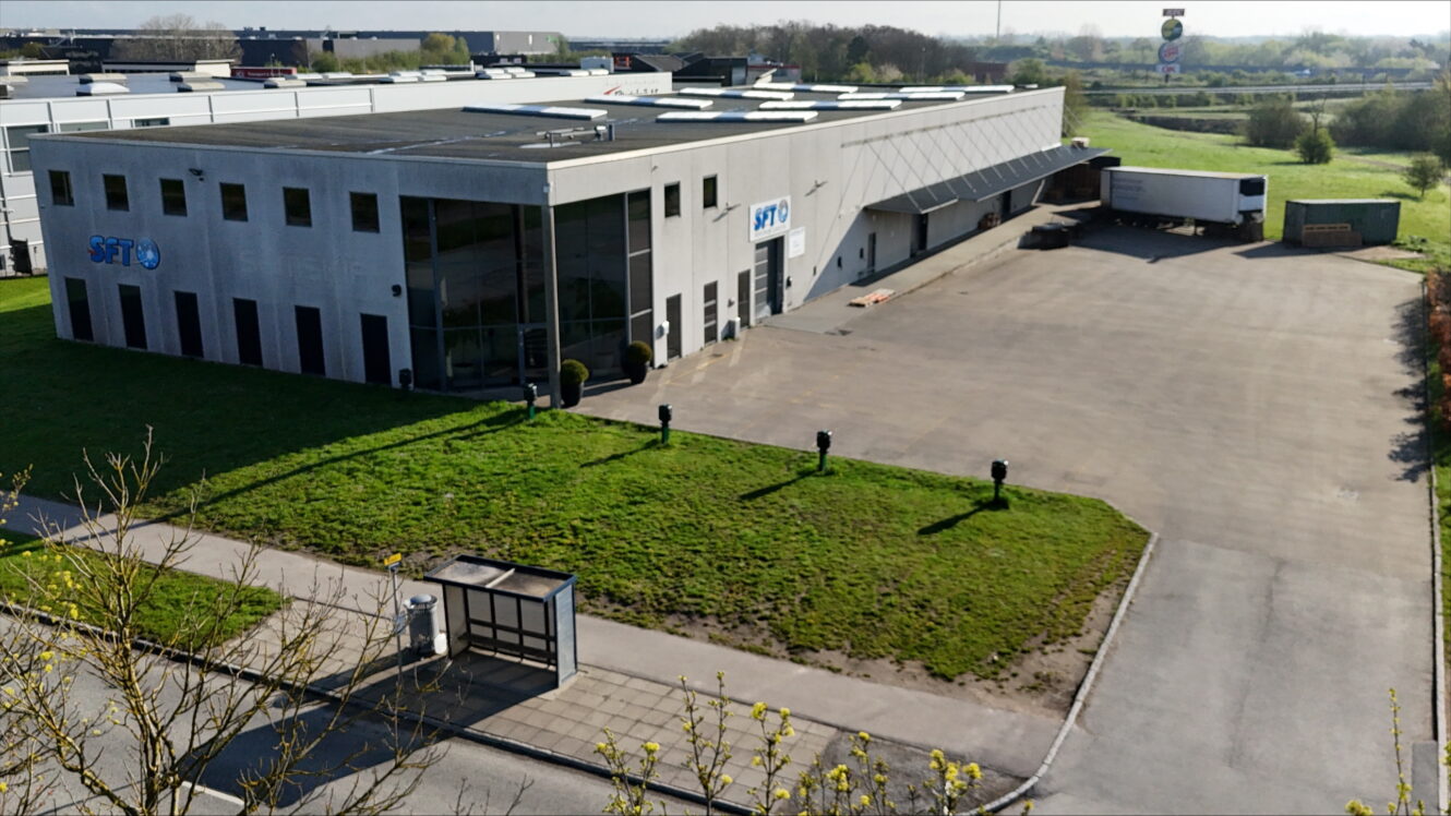 Et stort industrilager med glasvinduer og læsseramper, der støder op til en asfalteret plads med en parkeret sættevogn, omgivet af græs og en vej.