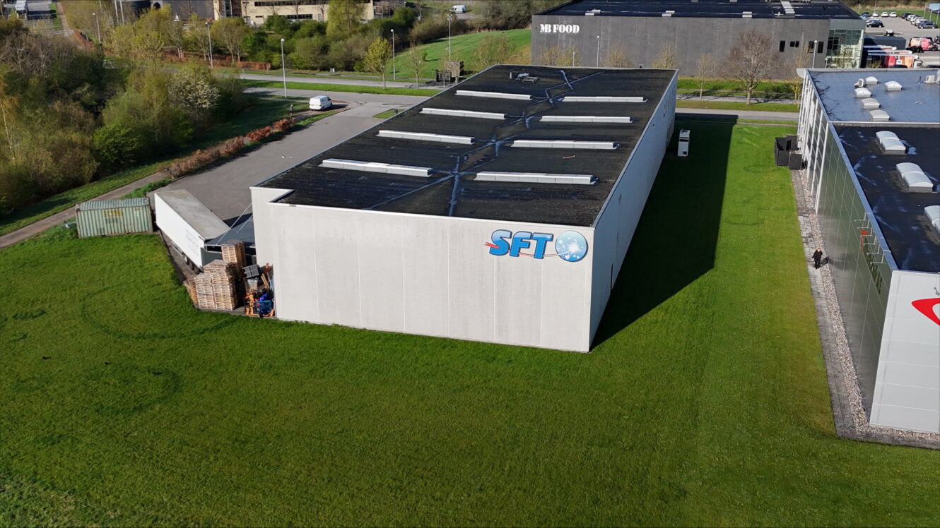 En stor hvid industribygning med bogstaverne SFT og et blåt globuslogo på siden, omgivet af græs og ved siden af andre erhvervsbygninger.