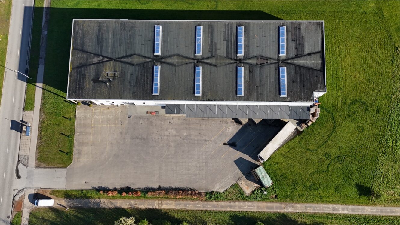 Luftfoto af et rektangulært industrilager med mørkt tag, ovenlysvinduer, læsseramper og omkringliggende asfalterede og græsklædte områder.