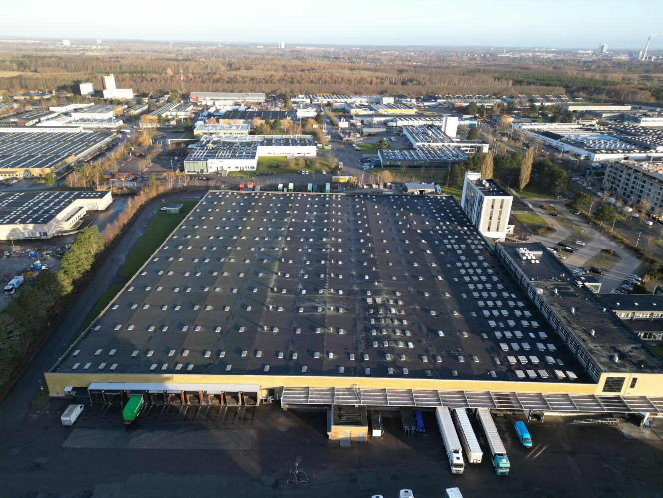 Luftfoto af et stort industrilager med fladt tag, læsseramper og flere lastbiler parkeret foran, omgivet af andre erhvervsbygninger.