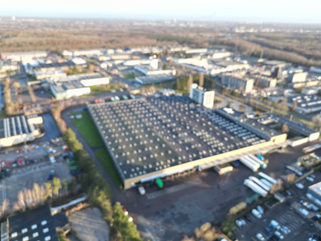 Luftfoto af et industriområde med en stor lagerbygning med fladt tag, omgivet af veje, parkeringspladser, lastbiler og andre bygninger.