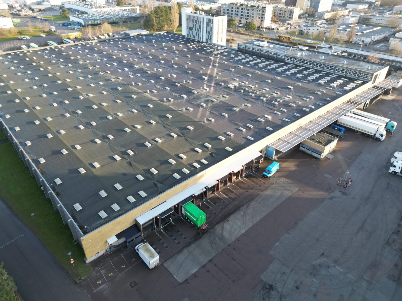 Et stort industrilager med flere læsseramper, flere lastbiler parkeret ved kajerne og den omkringliggende byinfrastruktur synlig i baggrunden.