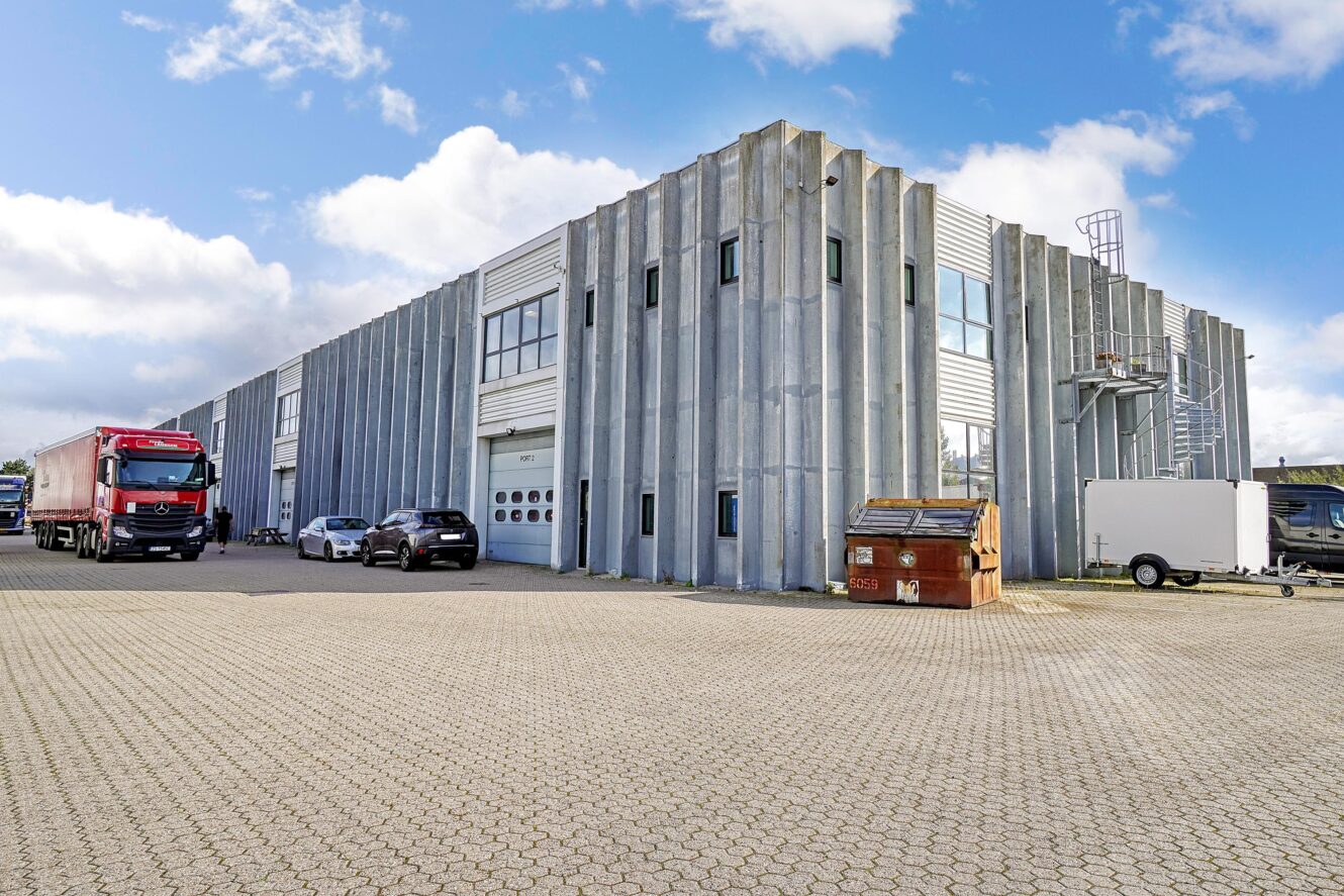 En moderne industribygning med lodrette betonelementer, parkerede biler, en lastbil, en container og en trailer under en delvis overskyet himmel.