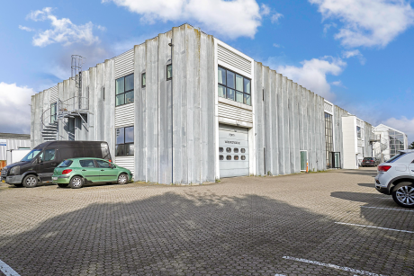 En moderne industribygning med bølgepapbeklædning, store vinduer og en vindeltrappe, omgivet af parkerede biler på en asfalteret plads under en delvis overskyet himmel.