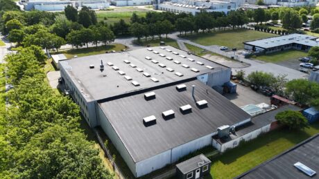 En stor industribygning med fladt tag og flere ovenlysvinduer og ventilationsenheder, omgivet af træer og andre kommercielle strukturer.