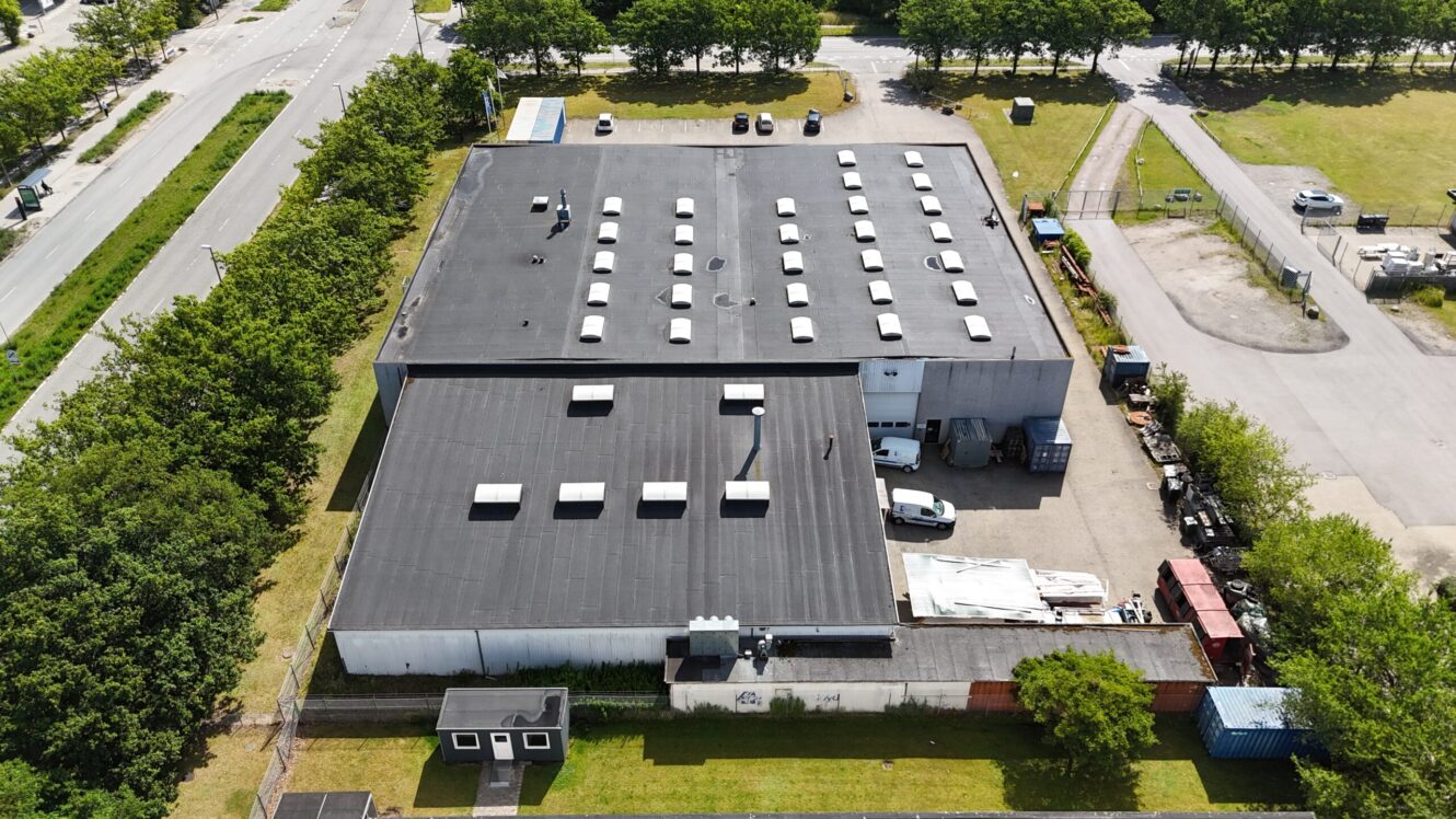 Luftfoto af en stor industribygning med fladt tag, mange ovenlysvinduer, parkerede biler og omkringliggende grønne områder og veje.