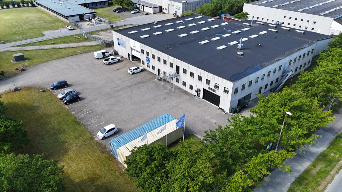 Luftfoto af en toetagers industribygning med flere parkerede biler og varevogne på parkeringspladsen, omgivet af græs og træer.