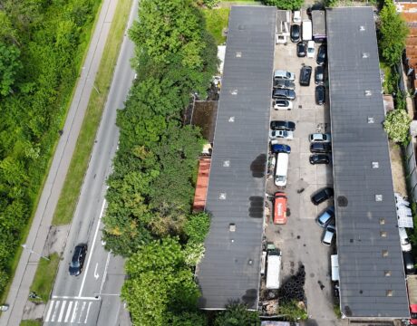 Luftfoto af en rektangulær parkeringsplads mellem to lange bygninger med flere parkerede biler ved siden af en vej med træer og en fodgængerovergang.