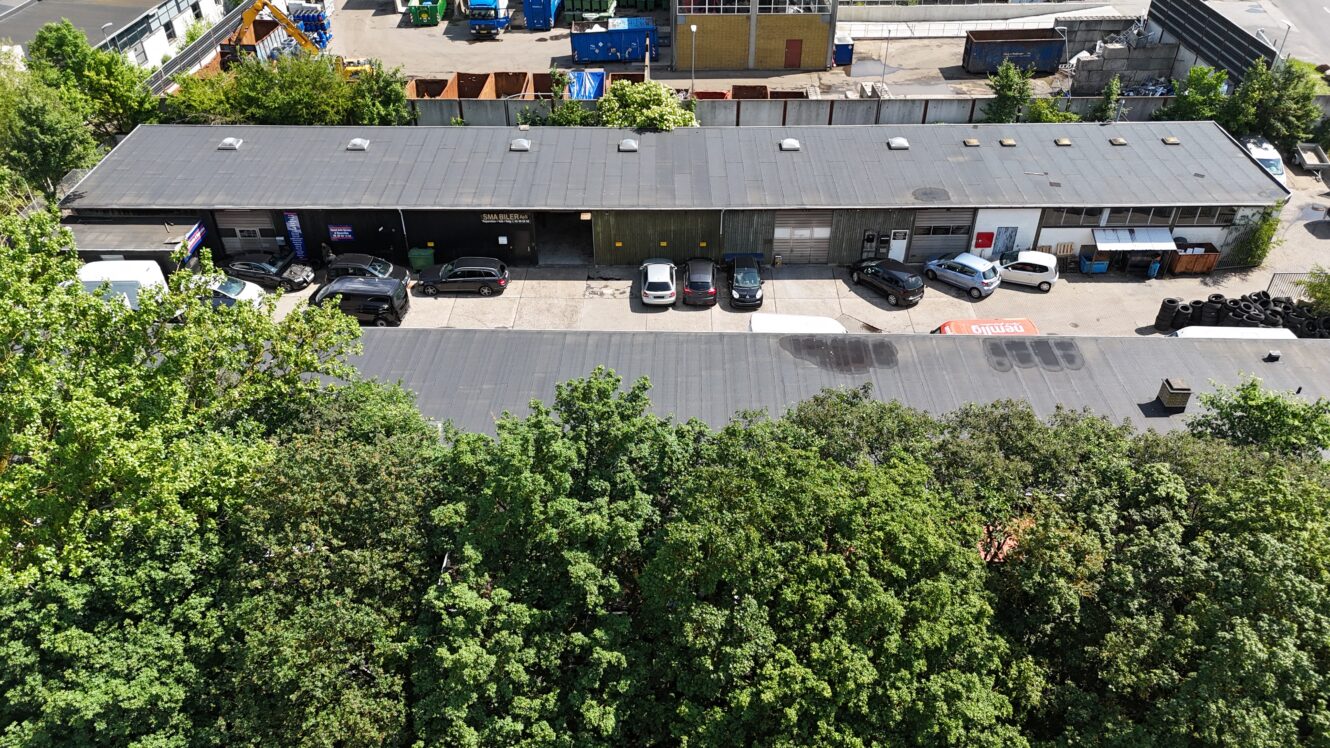 Luftfoto af en lang, enetages industribygning med parkerede biler foran og tætte, grønne træer i forgrunden.