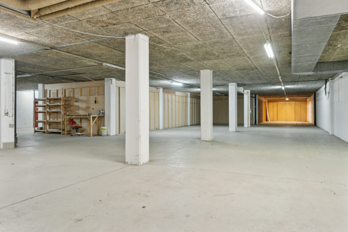 Stor, tom industrikælder med betongulv, hvide søjler, lysstofrør og træhylder langs den ene væg.