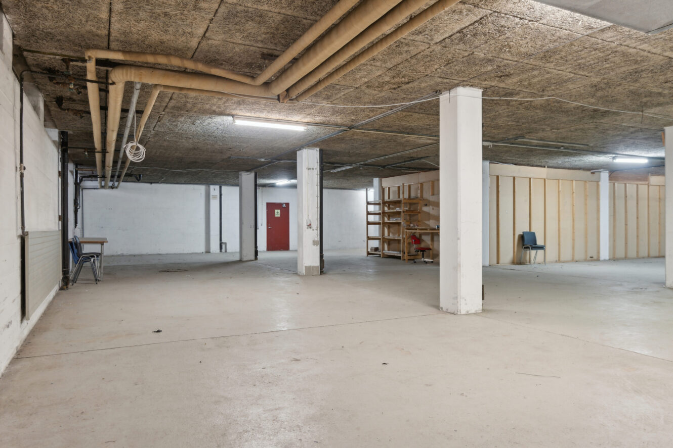 Rummelig, tom kælder med betongulv, synlige rør i loftet, lysstofrør og et par spredte stole og hylder langs væggene.