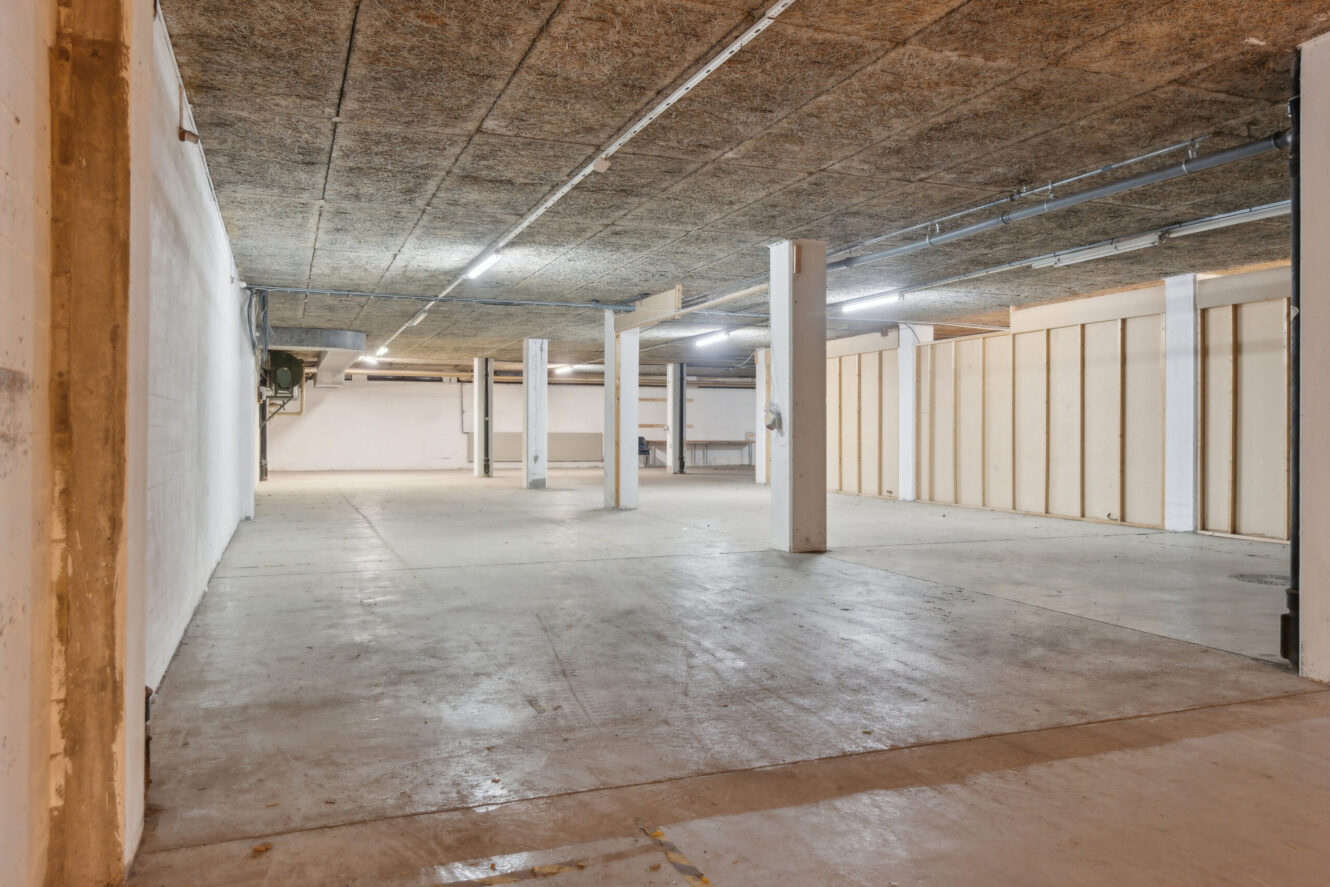 Rummelig, tom kælder med betongulv, synligt loft, søjler, lysstofrør og ufærdige vægge.