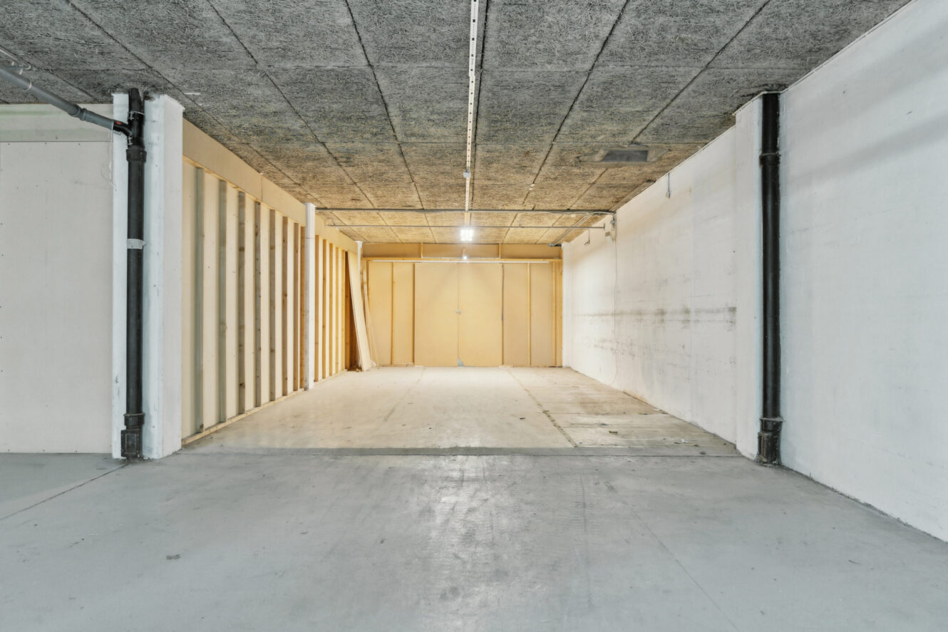 Tom garage i industristil med betongulv, hvide vægge, synlige loftspaneler og en lukket lysebrun dobbeltdør på bagsiden, oplyst af loftslamper.