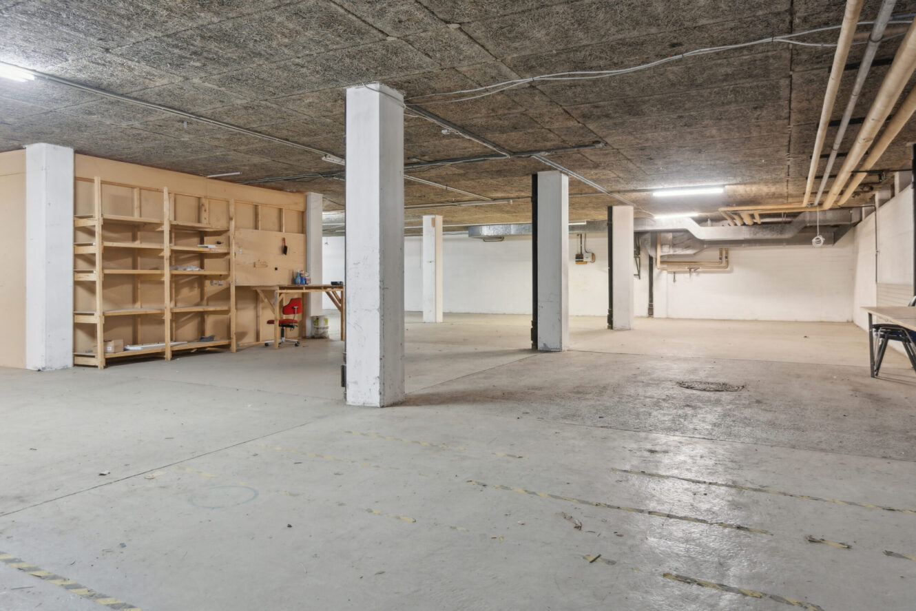 Rummelig, ufærdig kælder med betongulv, synligt loft, bærende søjler, reoler, lysstofrør og minimalt med møbler.