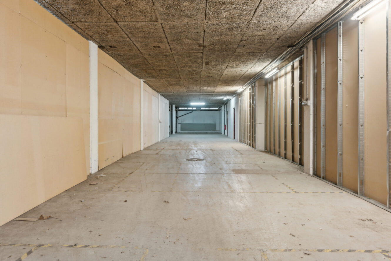 Tom, ufærdig korridor med blottet loft, umalede gipsvægge og betongulv, oplyst af lysstofrør langs den ene side.