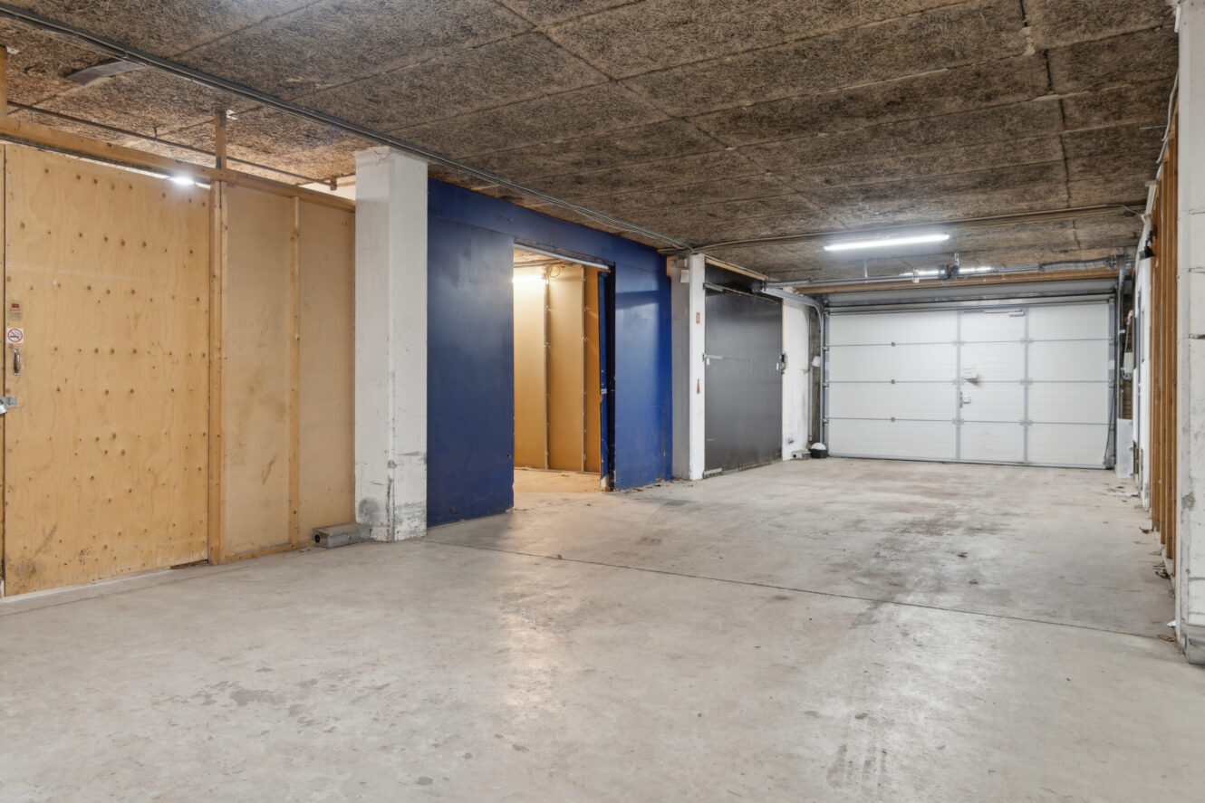 Rummelig indendørs garage med betongulv, opbevaringsenheder i træ til venstre, blåmalet væg i midten og en stor hvid rulleport til højre.