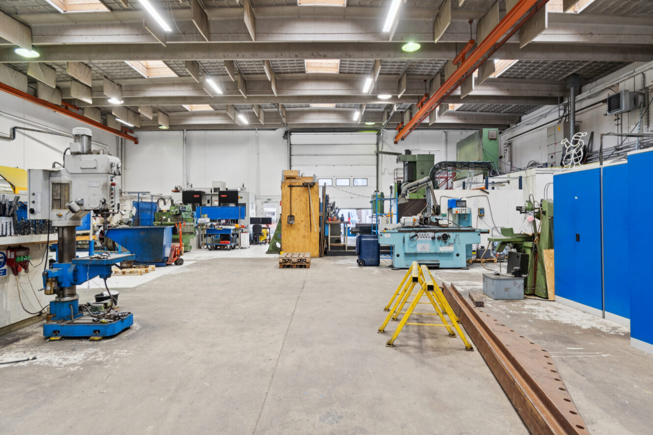 Rummeligt industrielt værksted med forskellige store maskiner, arbejdsborde, ovenlys og sikkerhedsbarrierer på et betongulv.