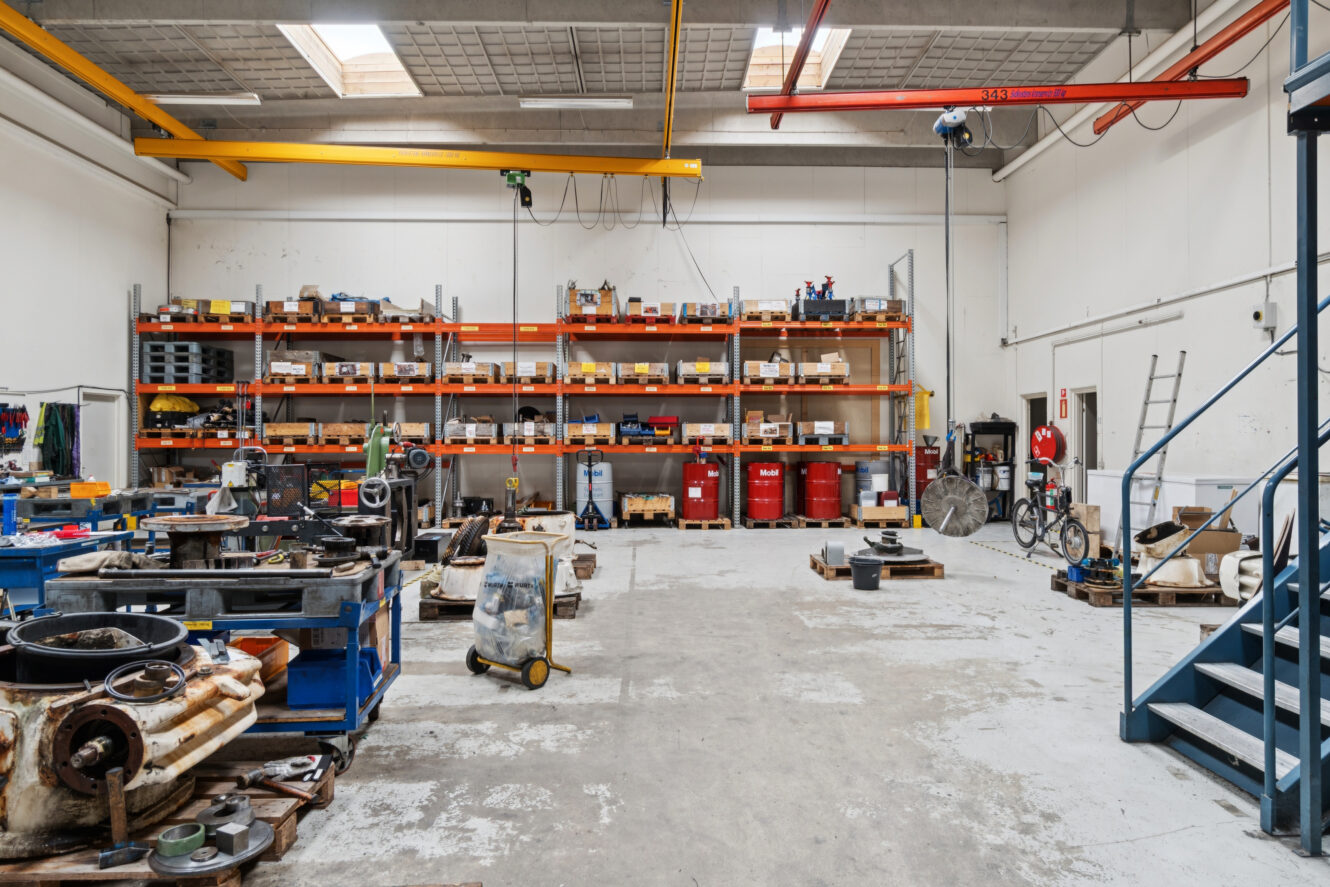 Rummeligt industrielt værksted med hylder med udstyr og værktøj, forskellige maskindele på gulvet og metaltrapper, der fører op til et øvre niveau.