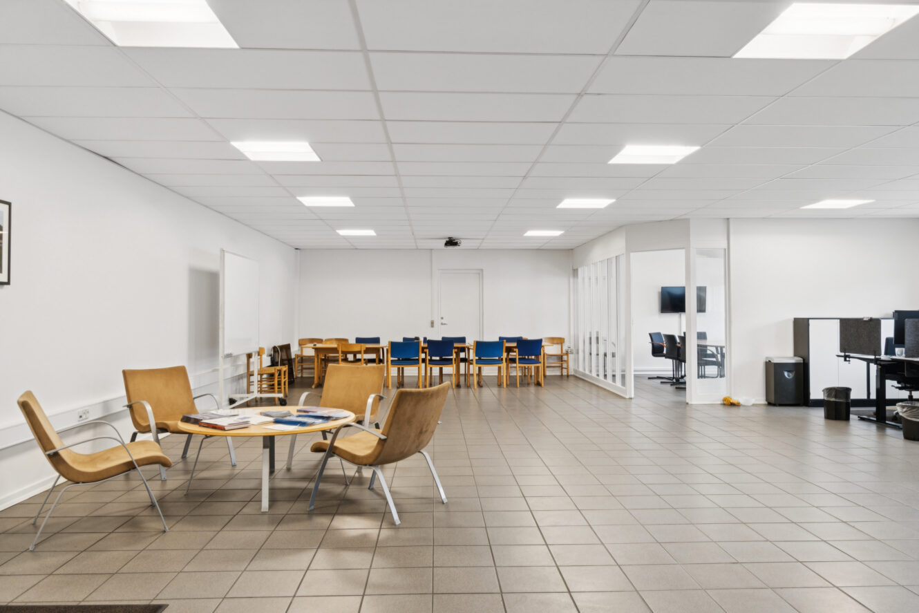 Et rummeligt, lyst lokale med klinkegulv, moderne stole omkring et lille bord og rækker af stablede træstole mod bagvæggen. Kontorerne er synlige gennem glasskillevægge.